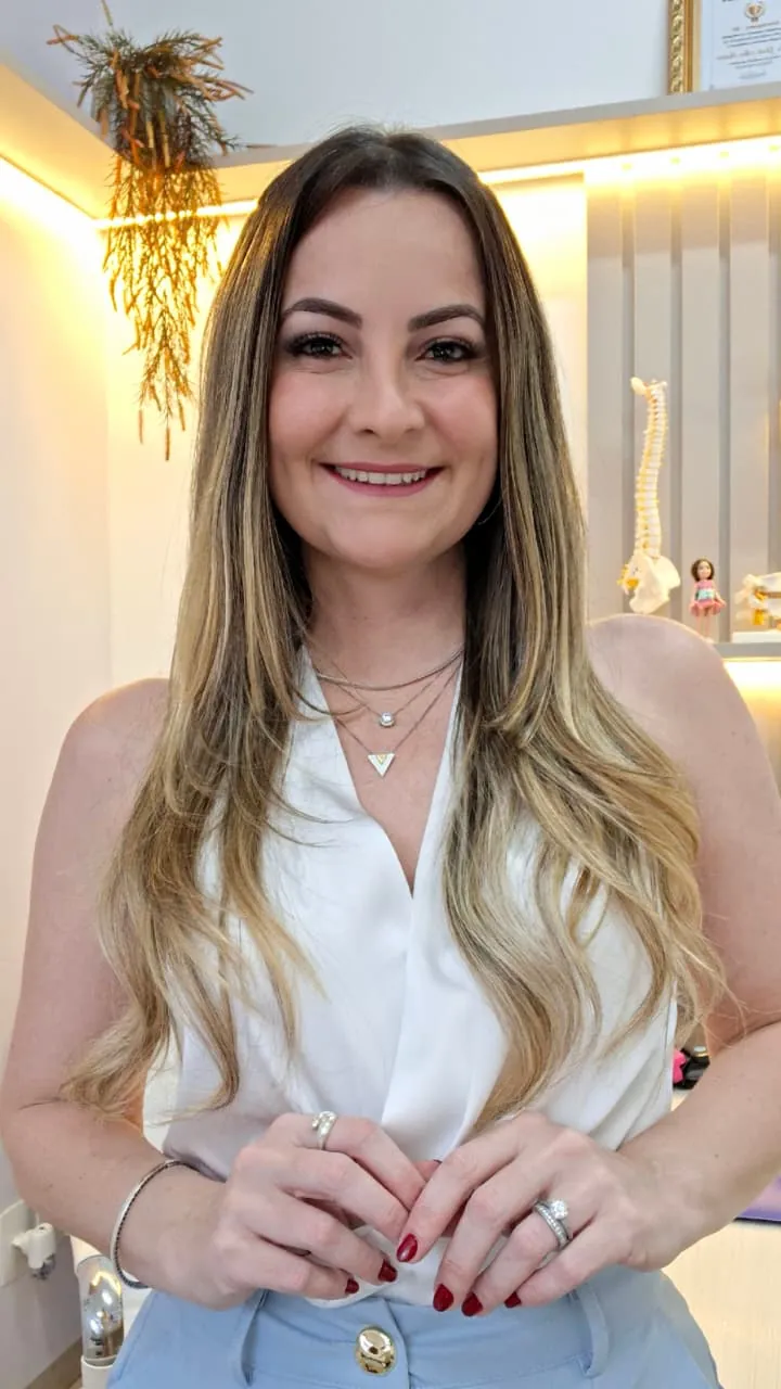 Dra. Gisela sorrindo, de blusa branca, no consultório com iluminação quente e um modelo de coluna ao fundo
