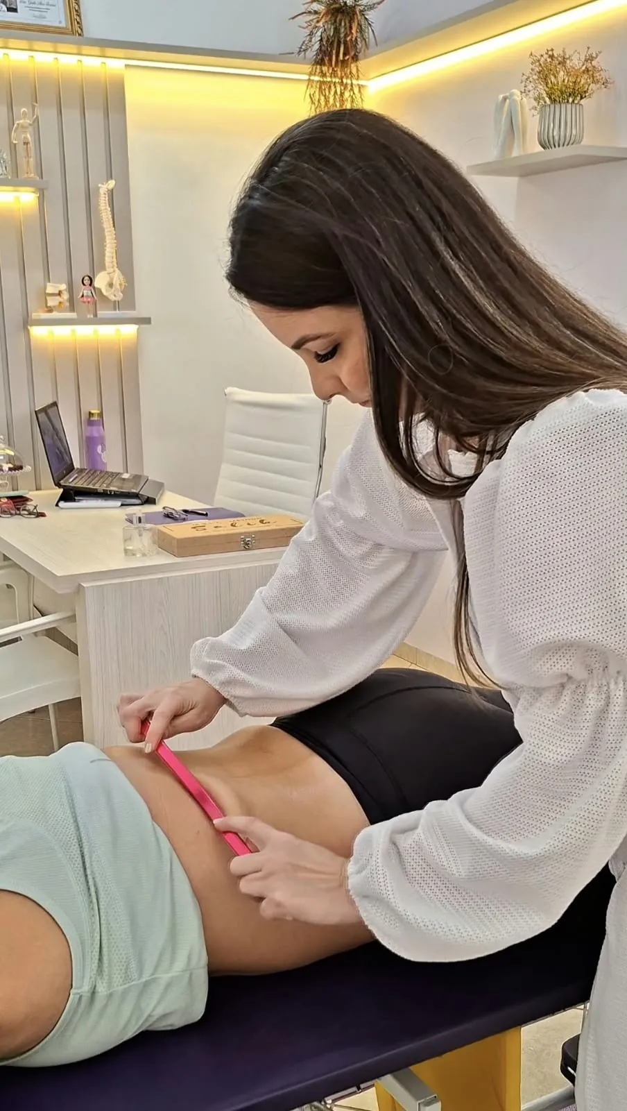 Dra. Gisela de blusa branca usando uma régua rosa para avaliar a região lombar de uma paciente deitada na maca