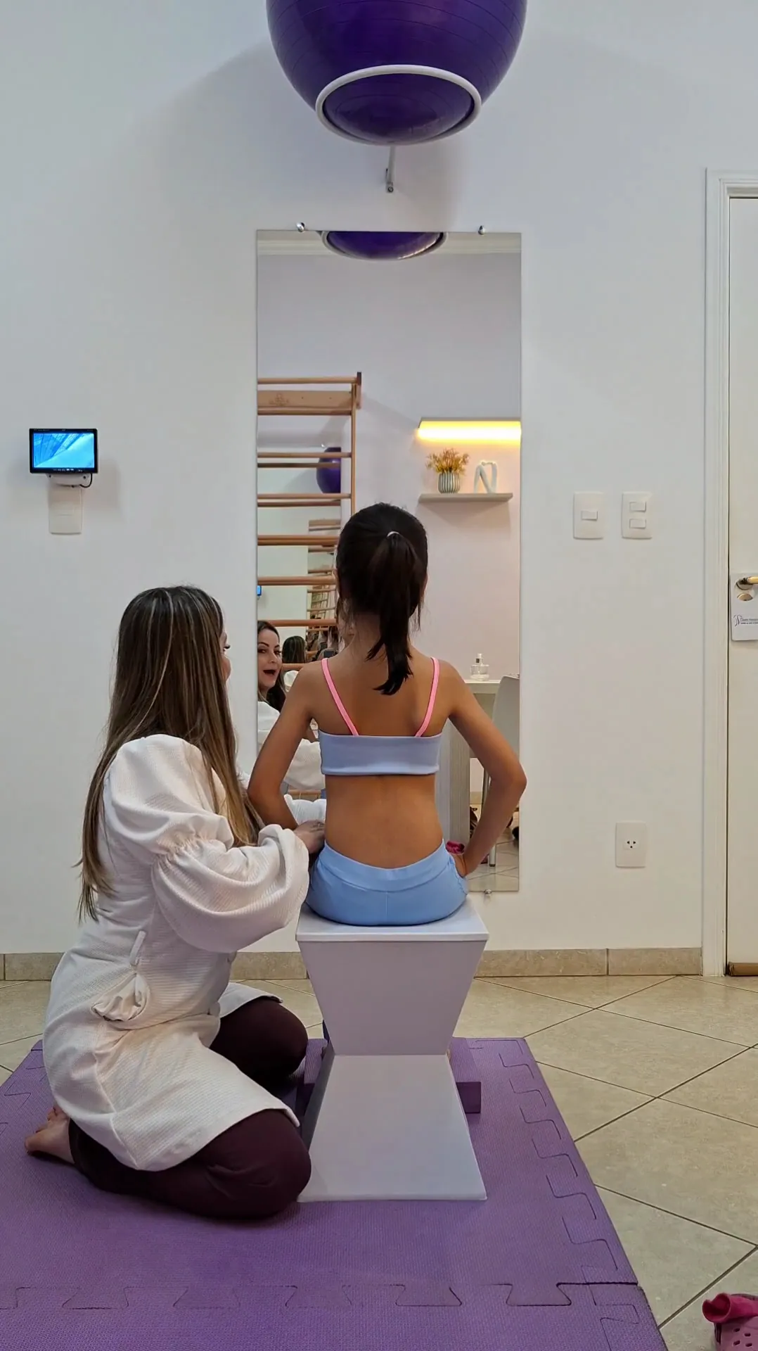 Dra. Gisela e uma menina de costas sentada diante do espelho, durante a avaliação postural